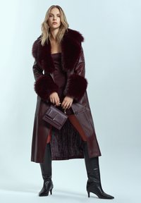 Cappotto in pelle bordeaux con dettagli in pelliccia sintetica, maniche lunghe e vestibilità sartoriale. Abbinato a stivali neri sopra il ginocchio e una borsa coordinata.