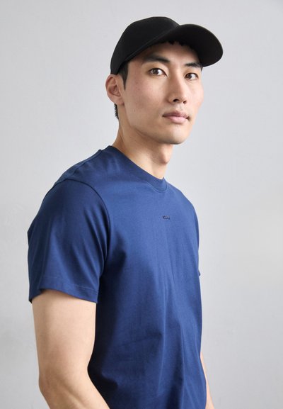 T-shirt in cotone blu navy con colletto rotondo e maniche corte, dotata di un piccolo logo sul davanti. Un berretto da baseball nero completa il look.