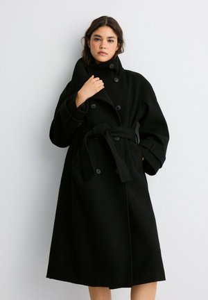 Femme aux cheveux foncés portant un long manteau noir ceinturé avec de gros boutons, debout devant un fond gris clair uni.