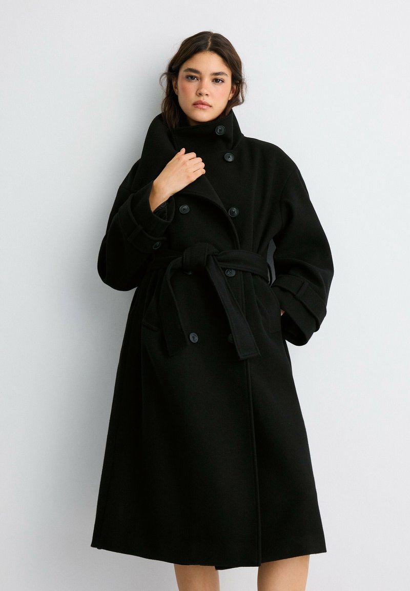 Femme aux cheveux foncés portant un long manteau noir ceinturé avec de gros boutons, debout devant un fond gris clair uni.