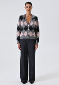 Schwarze, graue und rosa Argyle-musterte Strickjacke mit V-Ausschnitt, Knopfverschluss, weichem Material und geripptem Saum, kombiniert mit dunklen, weit geschnittenen Hosen.
