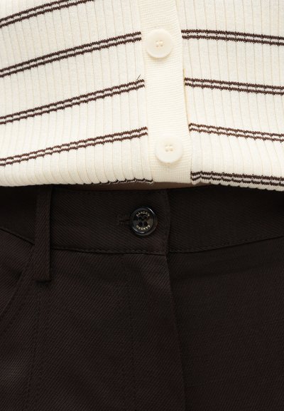 Κοντινό πλάνο εκρού ριμπ πλεκτού τοπ με καφέ ρίγες και κουμπιά, συνδυασμένο με ψηλόμεσο μαύρο παντελόνι στερεωμένο με μαύρο κουμπί.