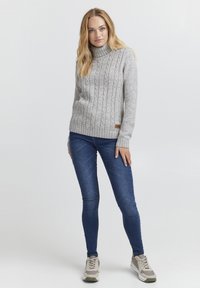 Lys grå kabelstrikket genser med høy krage, med ribbede mansjetter og nederkant. Kombinert med mørk blå skinny jeans og grå sneakers.