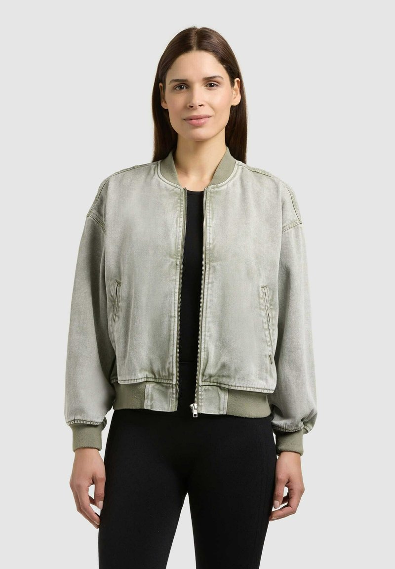 Femme aux cheveux bruns raides portant une veste bomber gris clair par-dessus un haut noir, debout devant un fond uni gris clair.