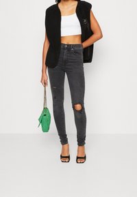 Mujer que lleva un chaleco de forro polar sin mangas negro, una camiseta corta blanca, jeans ajustados negros rasgados, tacones abiertos negros y sostiene un bolso de mimbre verde.