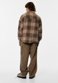 Rjava karo karo flanelska srajca z ohlapnim krojem, dolgimi rokavi in ukrivljenim robom, kombinirana z ohlapnimi bež cargo hlačami in priložnostno obutvijo.