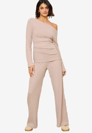 Béžový žebrovaný dvoudílný outfit sestávající z asymetrického svrchního dílu s dlouhým rukávem a širokých kalhot. Měkká textura, upnutý design zvýrazňuje siluetu.