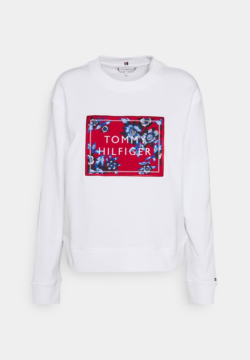 Sweat-shirt blanc à manches longues avec un patch rectangulaire rouge présentant des motifs floraux bleus et le texte "Tommy Hilfiger" en lettres blanches.