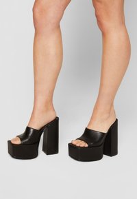 Talons à plateforme en cuir noir à bout ouvert avec des talons blocs épais et hauts portés sur pieds et jambes nus sur un fond uni.