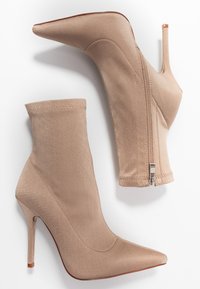 Par de botines de tacón alto en punta color beige con cremalleras laterales, fabricados en un tejido suave, exhibidos sobre un fondo blanco.