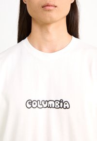 Camiseta de algodón blanca con un logo contorneado en negro que dice "COLUMBIA" en una fuente redondeada, con cuello redondo y textura suave.