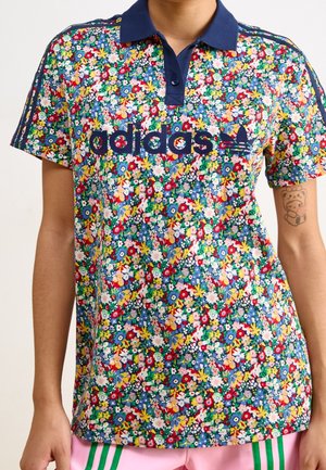 Osoba nosząca kolorową kwiatową koszulkę polo Adidas z granatowym kołnierzykiem i logo, w połączeniu z różowymi szortami z zielonymi paskami.