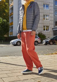 Person står på fortovet iført en grå pufferjakke, en gul hættetrøje, koralfarvede bukser og farverige sneakers i et bymiljø.