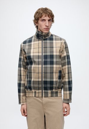 Jonge man met kort krullend haar draagt een beige, marineblauw en grijs geruit jack met rits en beige broek, staand tegen een effen witte achtergrond.
