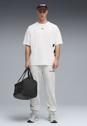 Wit T-shirt met korte mouwen en zwart logo, grijze joggingbroek met logo, en zwarte reistas. Grijze sneakers met groene accenten.