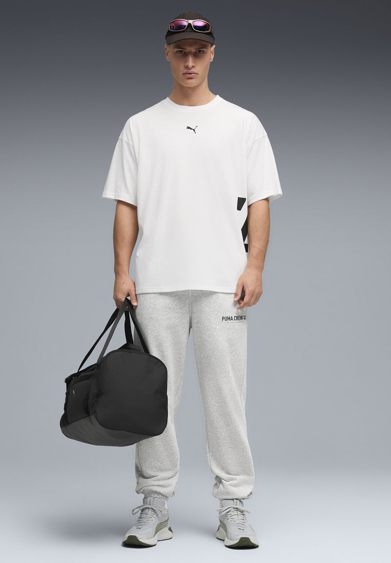 Bela majica s kratkimi rokavi s črnim logotipom, sive športne hlače z logotipom in črna torba za športno opremo. Sivi športni čevlji z zelenimi poudarki.