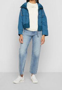 Zkrácená modrá péřová bunda s několika kapsami, zkombinovaná s světle béžovým tričkem a světle modrými džíny rovného střihu. Bílé tenisky.