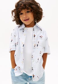 Νεαρό παιδί με σγουρά μαλλιά, φορούσε ανοιχτό γαλάζιο ριγέ πουκάμισο με στάμπες καλαθοσφαιριστών, λευκό μπλουζάκι και ανοιχτό γαλάζιο τζιν, με τα χέρια στις τσέπες.