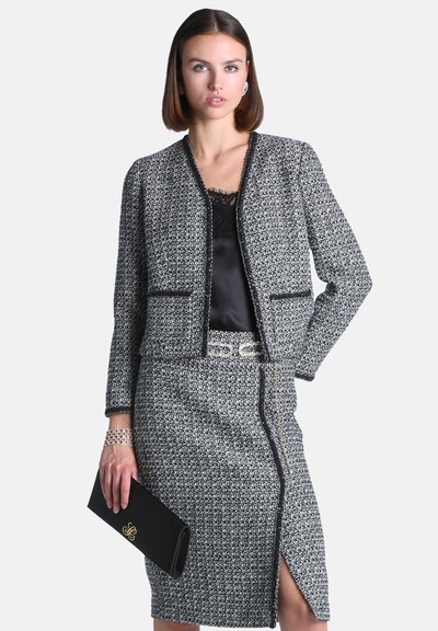 Femme en ensemble veste et jupe en tweed noir et blanc, tenant une pochette noire et portant un haut en dentelle noire en dessous.