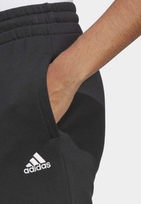 Fekete sportnadrág texturált anyagból, rugalmas derékpánttal és oldalzsebbel. Bal csípőn egy fehér Adidas logóval.