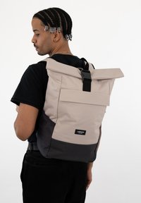 Roll-top backpack in beige and dark gray. Made of durable fabric, featuring a front flap, adjustable straps, and a prominent brand label.