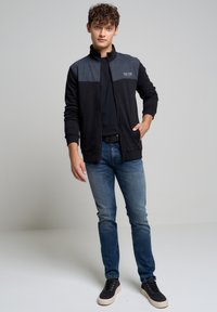 Giacca zip-up nera e grigia con colletto alto, parte superiore texturizzata e tasche laterali, abbinata a jeans blu e scarpe nere.