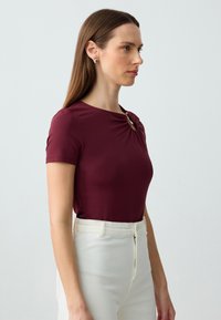 Burgundowa bluzka z krótkim rękawem i okrągłym dekoltem, z przekręconym detalem z przodu, z złotym akcentem. Połączona z białymi spodniami o wysokim stanie.
