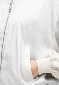 Χέρι στην τσέπη ενός λευκού σατέν μπουφάν τύπου bomber με διακριτικό σχέδιο, ασημένιο φερμουάρ και ριμπ μανσέτα στο μανίκι.