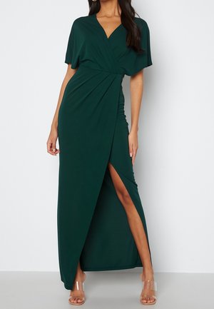 Woman wearing a dark green wrap-style maxi dress with short sleeves and a thigh-high slit, paired with clear strap heels.
