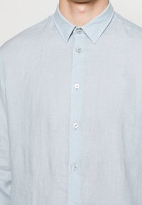 Uomo che indossa una camicia in lino azzurro chiaro con colletto a punta, primo bottone allacciato e tre bottoni visibili lungo la parte anteriore.