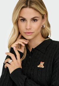 Juodas megztinis su kabelių raštu ir apykakle, dešinėje krūtinės pusėje puikuojasi išsiuvinėtas meškiuko dizainas. Modelis vilki sidabrinius žiedus.