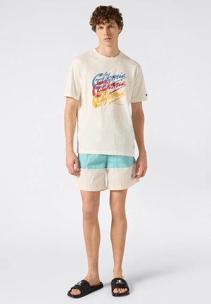 Młody mężczyzna w kremowej koszulce z nadrukiem "California", niebieskich i kremowych szortach oraz czarnych klapkach Champion, stojący na jednolitym białym tle.