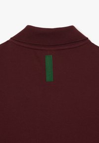 Gros plan d'un col en maille bordeaux avec une étiquette en tissu rectangulaire vert foncé cousue verticalement sous l'encolure arrière.