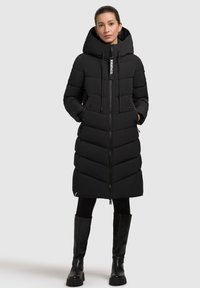 Abrigo acolchado negro con capucha, cierre de cremallera y patrón de chevrón. Presenta bolsillos laterales y cordones ajustables. Combinado con botas negras.