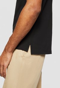 T-shirt noir à manches courtes avec fente sur le côté, porté avec un pantalon beige clair. Le tissu semble lisse avec une finition légèrement texturée.