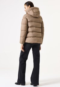 Chaqueta de plumas marrón con capucha, que presenta secciones acolchadas y un acabado ligeramente brillante. Llevada con jeans oscuros de campana y calzado negro.