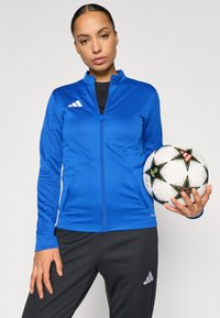 Μπλε αθλητικό μπουφάν με φερμουάρ και λευκές λεπτομέρειες, σε συνδυασμό με μαύρο αθλητικό παντελόνι. Το μοντέλο κρατά μια ποδοσφαιρική μπάλα με γεωμετρικό σχέδιο.