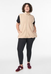 Beige fleeced vest met hoge kraag, front rits en twee zijzakken, gecombineerd met zwarte leggings en bordeauxrode sneakers.