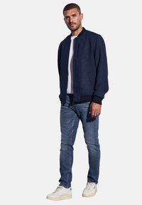 Hombre con chaqueta bomber azul marino, camiseta blanca, jeans azules y zapatillas blancas de pie contra un fondo blanco.