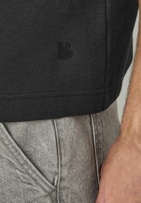 T-shirt en coton noir avec une légère texture, arborant un petit logo noir sur le côté. Assorti à un pantalon en denim gris clair.