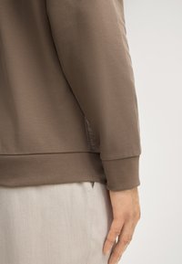 Felpa marrone in cotone con orlo e polsini a costine. Il tessuto appare liscio, aderente sopra pantaloni chiari.