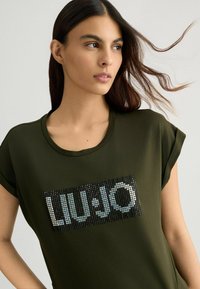 T-shirt a maniche corte verde oliva con logo decorato con paillettes nere sul davanti. Il tessuto è morbido e liscio con scollatura rotonda.