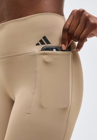 Χέρι τοποθετεί ένα smartphone στην πλαϊνή τσέπη μπεζ κολαν Adidas, με κοντινό πλάνο στο ισχίο και το λογότυπο.