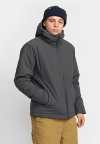 Chaqueta aislante gris con un alto cuello, cremallera frontal y bolsillos laterales. Combinada con un gorro azul oscuro. Material suave y corte relajado.