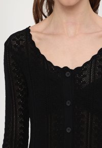 Čierny pletený kardigán s vlnkovým výstrihom, ktorý má textúrovaný vzor a päť predných gombíkov.