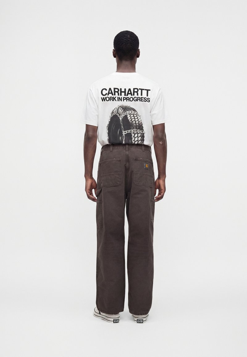 Mann steht mit dem Rücken zur Kamera, trägt ein weißes Carhartt-T-Shirt mit Reifen-Grafik und braune, weit geschnittene Hosen mit mehreren Gesäßtaschen.