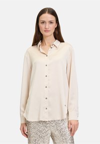 Helle beige Seidenbluse mit Kragen, Knopfleiste und langen Ärmeln. Sie hat eine glatte Textur und einen subtil abgerundeten Saum.