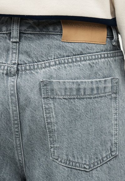 Κοντινό πλάνο του πίσω τσέπης σε ανοιχτό γαλάζιο τζιν με καφέ ετικέτα Maison Kitsuné και μέρος από ένα λευκό και ναυτικό πουκάμισο.