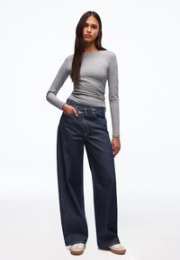 Šedé dlouhé přiléhavé tričko ve spojení s volnějšími tmavě modrými džíny s vysokým pasem a viditelným prošitím, nošené s béžovými teniskami.