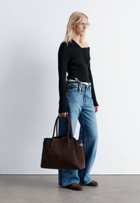 Braune Wildleder-Tasche mit zwei Griffen, rechteckiger Form und einer Detailnaht. Getragen mit einem schwarzen gerippten Pullover und blauen weit geschnittenen Jeans.
